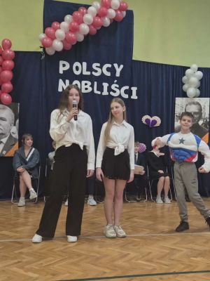 Dziewczęta z klasy VIb &ndash; Zosia i Julia &ndash; recytują wiersz Wisławy Szymborskiej &bdquo;Konkurs piękności męskiej&rdquo;, podczas gdy chłopcy Tymon i Janek zabawnie pozują, prężąc swoje mięśnie, nawiązując do treści wiersza i tworząc humorystyczną scenę.