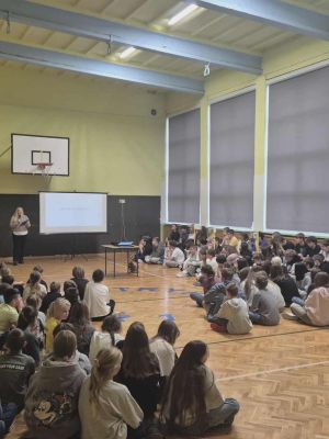 Wypełniona uczniami sala gimnastyczna, gdzie nauczycielka prowadzi apel na tle ekranu.