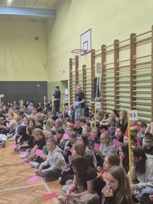 Duża grupa uczni&oacute;w siedzi na podłodze sali gimnastycznej podczas szkolnego apelu, trzymając w rękach kolorowe kartki.