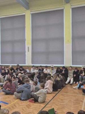 Duża grupa uczni&oacute;w siedzi w rzędach przy zasłoniętych oknach, czekając na dalszą część spotkania.